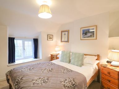 A bedroom with a double bed two bedside tables with lamps a window with blue curtains and framed pictures on the walls at River Cottage in Burton Bradstock