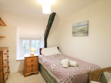 A bedroom with a single bed with floral bedding a wooden bedside table with a lamp a wooden chest of drawers a wicker chair and a window with a view at River Cottage in Burton Bradstock