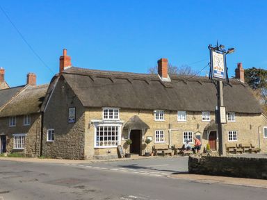 A stone building with a thatched roof and chimneys with people outside at Three Horseshoes in Burton Bradstock