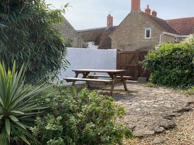 An outdoor patio with a wooden picnic table surrounded by plants and stone walls at River Cottage in Burton Bradstock