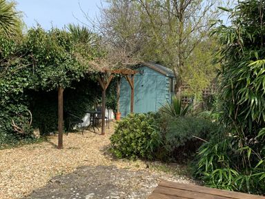 A garden with a wooden pergola seating area and a teal garden shed surrounded by plants and bushes at River Cottage in Burton Bradstock