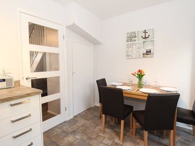 A dining area with a wooden table set for four people with plates glasses and a vase of flowers with a door and kitchen counter nearby at Rockpools Weymouth