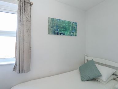 A small bedroom with a single bed white sheets blue pillow a window with curtains and a painting on the wall at Rockpools in Weymouth