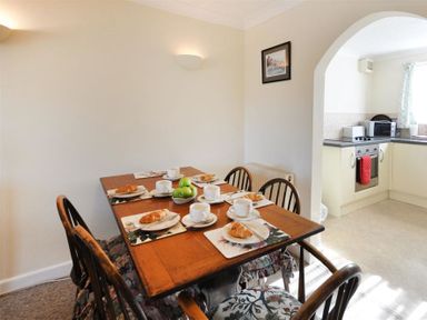 A dining area with a wooden table set for six with cups croissants and green apples next to a kitchen with appliances at Sanjernin in Swanage