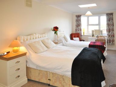 A bedroom with two beds a bedside table with a lamp a red chair a dressing table and window with floral curtains at Sanjernin in Swanage