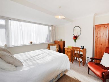 A bedroom with a bed, an armchair, wooden desk with a chair, wooden wardrobe, and a window with white curtains at Sanjernin Swanage