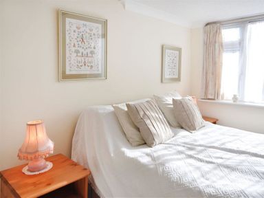 A bedroom with a bed covered in white bedding and beige pillows flanked by wooden nightstands with lamps and framed pictures on the wall at Sanjernin in Swanage