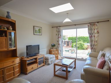 A living room with a sofa, coffee table, television on a wooden stand, floral curtains, and sliding glass doors opening to a garden at Sanjernin in Swanage