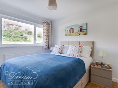 A bedroom with a bed with a blue blanket two pillows a bedside table with a lamp cups and books and a window with curtains at Seacliff in West Bay
