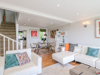 A living and dining area with white sofas and colorful cushions a wooden dining table with chairs a staircase and a shelving unit at Sea King House in Cove Castle