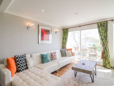 A living room with a white sectional sofa with colorful cushions a wooden coffee table green curtains and sliding glass doors at Sea King House in Cove Castle