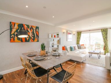 Dining table with plates and glasses next to a living room with white sofa and green curtains at Sea King House in Cove Castle