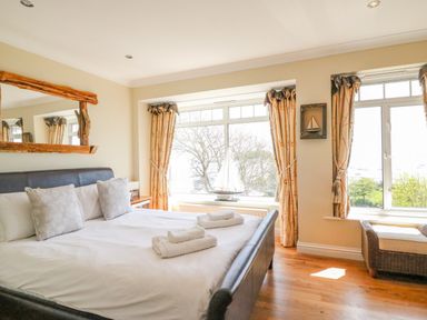 A bedroom with a bed, pillows, two towels, large windows with curtains, a wooden floor, and a wicker chair at Sea King House in Cove Castle