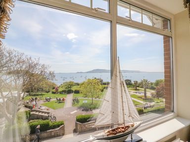 A window view of a park with people sitting and walking near the sea with boats sailing and a model sailboat on the windowsill at Sea King House in Cove Castle