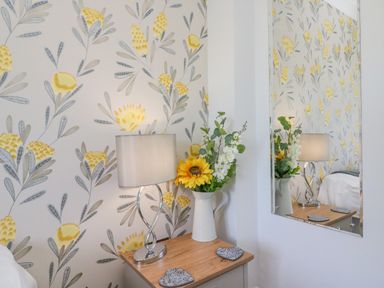 A bedside table with a lamp and vase of flowers next to a wall with floral wallpaper and a mirror reflecting the scene at Sea King House in Cove Castle
