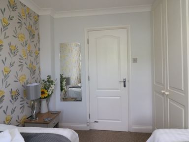 A bedroom with floral wallpaper a bedside table with a lamp and flowers a white door and built in wardrobes at Sea King House in Cove Castle