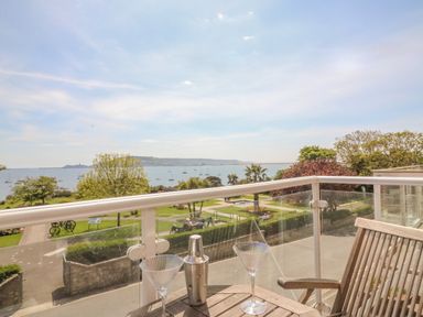 A balcony with a wooden table and chairs overlooking a park and a body of water with boats at Sea King House in Cove Castle