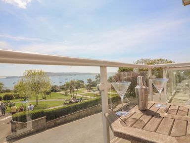A balcony with two empty cocktail glasses and a metal shaker overlooking a park with people and a sea with boats at Sea King House in Cove Castle