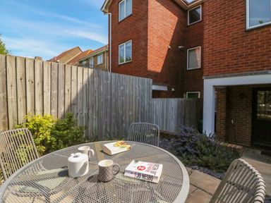 An outdoor patio with a round metal table, chairs, a teapot, mugs, and books next to a brick building and wooden fence at Sea King House in Cove Castle