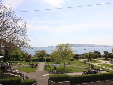 A park with people sitting on benches and tables near a body of water with boats and hills in the background at Sea King House in Cove Castle