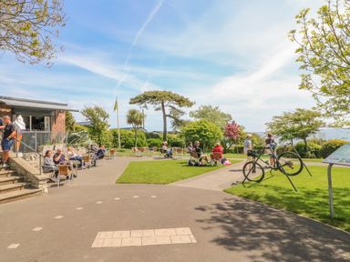 An outdoor seating area with people sitting on chairs near a brick building and a bicycle parked on a path at Sea King House in Cove Castle