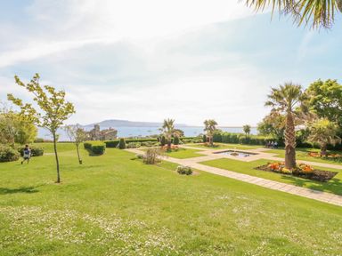 A garden with palm trees flower beds benches and a view of the sea at Sea King House in Cove Castle