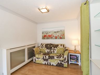 A small room with a floral patterned sofa a side table with a lamp and board games a wall painting and a green curtain at Sea Pebbles in Weymouth