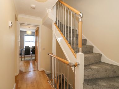 A hallway with wooden floor leading to a dining room and a staircase with carpeted steps and wooden handrails at Sea Pebbles in Weymouth