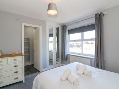 A bedroom with a bed with towels a window with curtains a chest of drawers and a door leading to a bathroom at Sea Pebbles in Weymouth