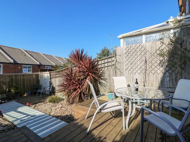 An outdoor wooden deck with a glass table and four chairs next to a fenced garden at Sea Pebbles in Weymouth