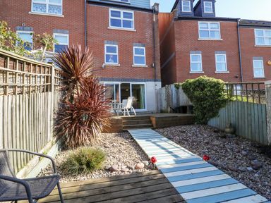 A backyard with a wooden pathway and patio furniture behind a brick townhouse at Sea Pebbles in Weymouth