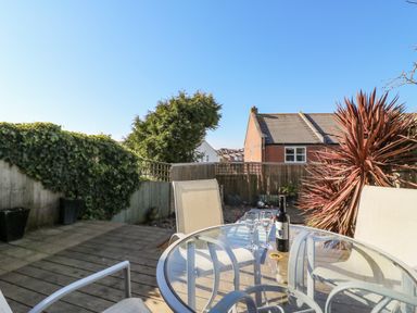 An outdoor patio with a glass table and chairs with a bottle of wine and two glasses surrounded by plants and a fence at Sea Pebbles in Weymouth