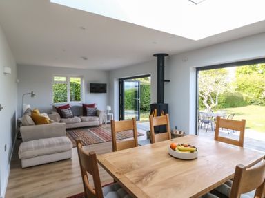 A dining table with chairs and a bowl of fruit in an open living room with sofas a wood stove and doors opening to a garden at Shipton Hill View in Shipton Gorge