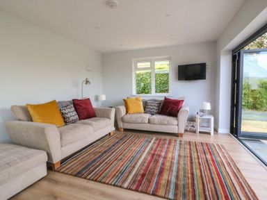 A living room with two beige sofas with cushions a striped rug a small white side table a floor lamp a wall-mounted TV and a glass door at Shipton Hill View in Shipton Gorge