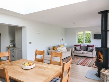 A dining table with chairs and fruit bowl next to a living room with sofas and a wood-burning stove at Shipton Hill View in Shipton Gorge
