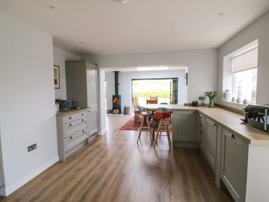 A kitchen with wooden floors and light gray cabinets leading to a dining area with a wood stove and large folding doors at Shipton Hill View in Shipton Gorge