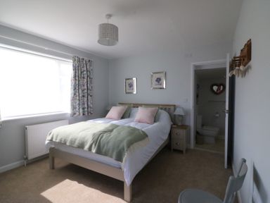A bedroom with a double bed, bedside tables with lamps, a window with floral curtains and an open door to a bathroom at Shipton Hill View in Shipton Gorge