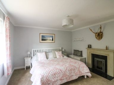 A bedroom with a bed with pink patterned bedding bedside tables lamps a fireplace and a wooden deer head at Shipton Hill View in Shipton Gorge