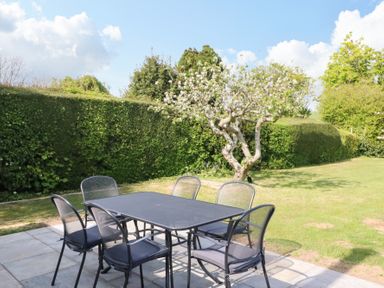 An outdoor patio with a black metal table and six chairs on a stone floor with green hedges and a blossoming tree in the background at Shipton Hill View in Shipton Gorge