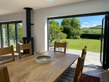 A dining area with a wooden table and chairs next to large open doors leading to a garden with patio furniture and greenery at Shipton Hill View in Shipton Gorge