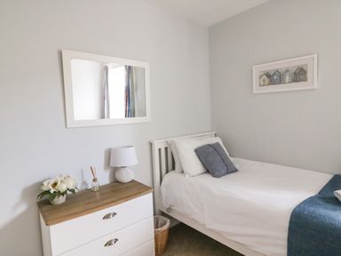 A bedroom with a white bed a wooden dresser with a lamp and flowers and a wall mirror at Shipton Hill View in Shipton Gorge