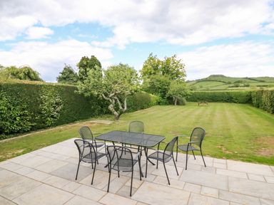 A patio with a metal table and six chairs overlooking a garden with grass trees and hedges at Shipton Hill View in Shipton Gorge