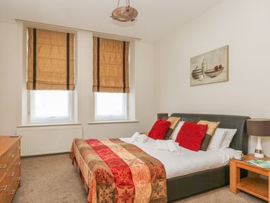A bedroom with a double bed featuring red and brown pillows and a patterned bedspread with two windows covered by brown Roman blinds and wooden furniture at Sea Star in Weymouth