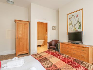 A bedroom with a wooden wardrobe a wooden dresser with a TV a leather chair and an open door to a bathroom at Sea Star in Weymouth