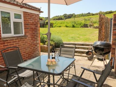 An outdoor patio with a glass table and chairs a bottle of wine and cupcakes on the table a barbecue grill and a grassy garden area at Studland Cottage in Studland