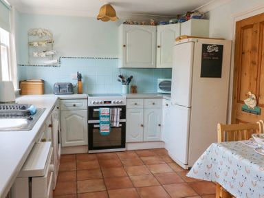 A kitchen with white cabinets a white refrigerator an oven with towels on the handle a wooden door and a table with a chicken patterned tablecloth at Studland Cottage in Studland