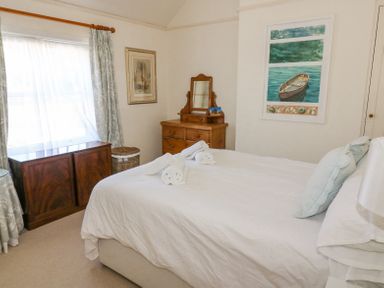 A bedroom with a bed made with white linens two light blue pillows a wooden dresser with a mirror a small cabinet a window with sheer curtains and a painting of a rowboat on the wall at Studland Cottage in Studland