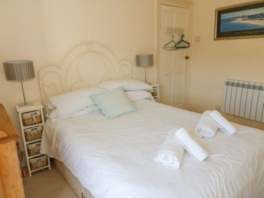 A bedroom with a double bed with white linens and folded towels a wooden dresser two bedside tables with lamps a door with hangers and a framed picture on the wall at Studland Cottage in Studland