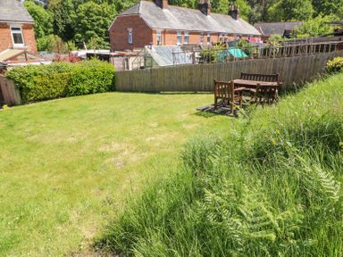 A backyard with green grass a wooden table and chairs a fence and brick houses in the background at Studland Cottage in Studland