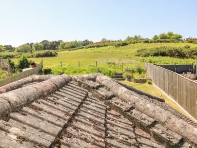 A view over an old tiled roof towards a green fenced garden and a grassy field with trees at Studland Cottage in Studland